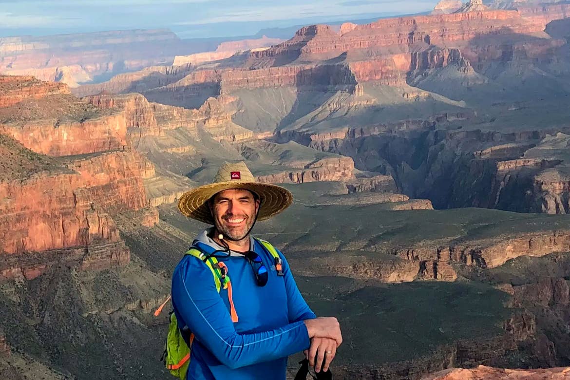 A caucasian male wearing a blue long-sleeve shirt and sunhat stands in front of the beautiful, mountainous Grand Canyon landscape.