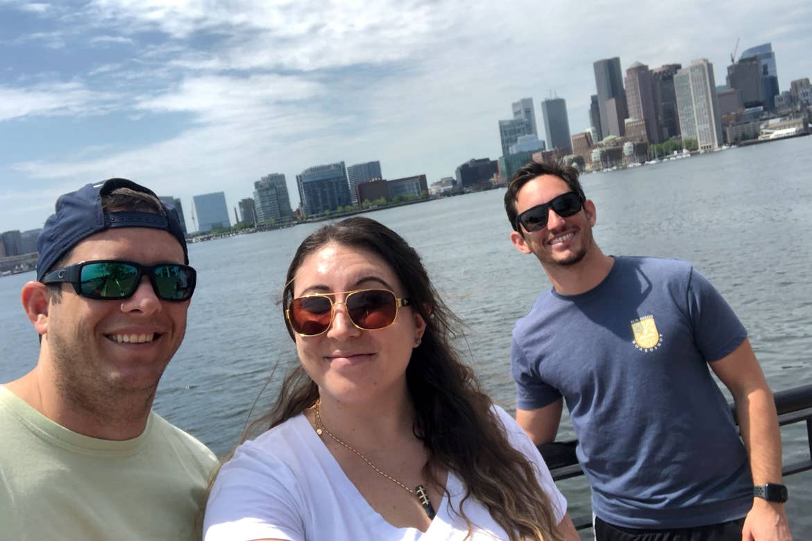 From left to right: A man, woman, and another man stand near a river in front of the Boston skyline.