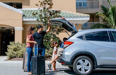 A woman, man and young boy and girl unpack a silver vehicle outside of a resort.