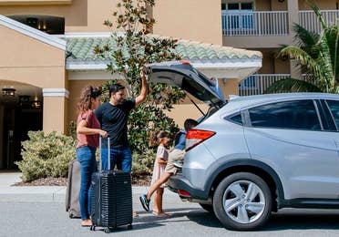 A woman, man and young boy and girl unpack a silver vehicle outside of a resort.