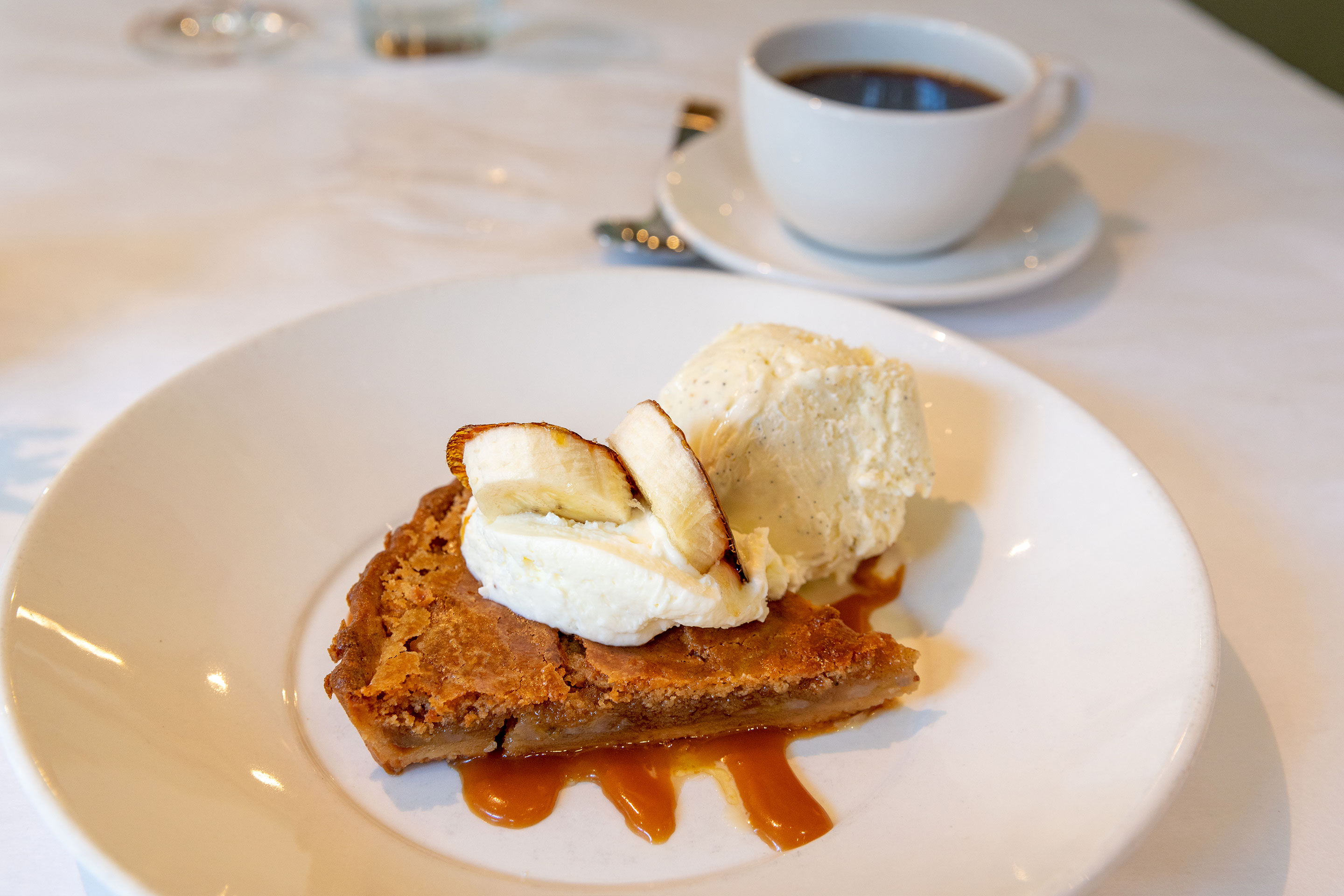 A white coffee mug and saucer placed next to a brown butter and banana tart on white plate.