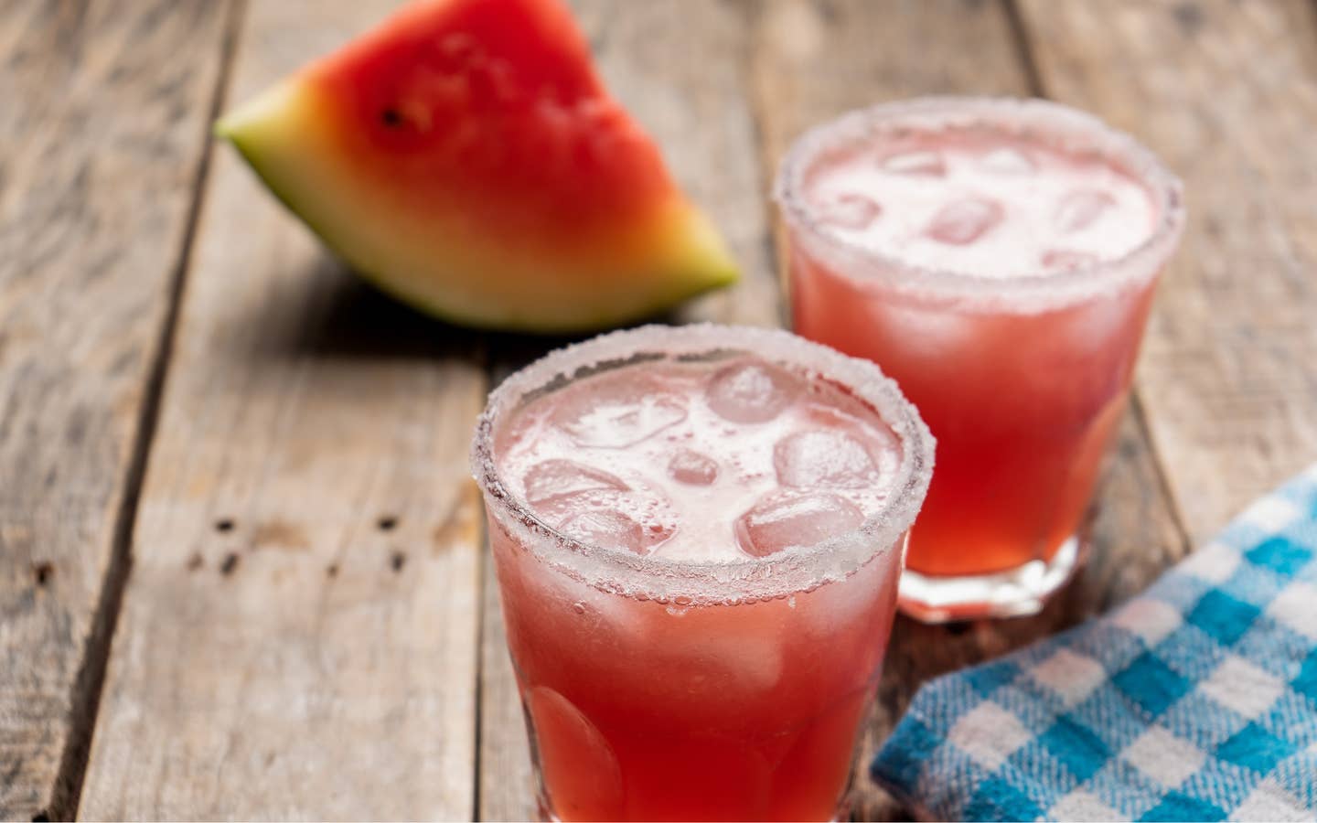 Two glasses of Melon Margaritas sit on a wooden plank table in front of a watermelon wedge.