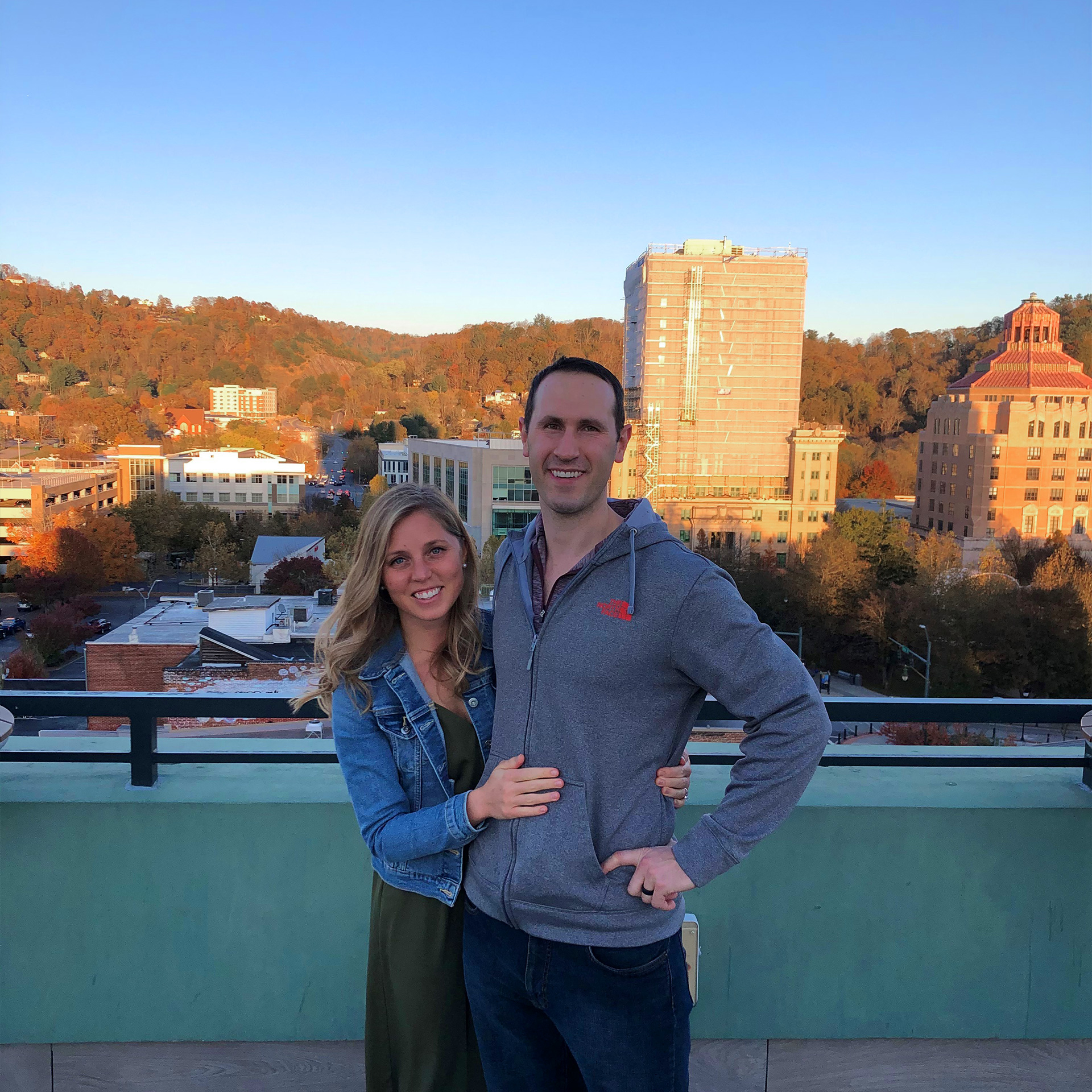 A blonde woman (left) wearing a denim jacket and green maxi dress hugs a man (right) wearing a grey zip-up hoodie on a rooftop bar overlooking the downtown area of Asheville, North Carolina.