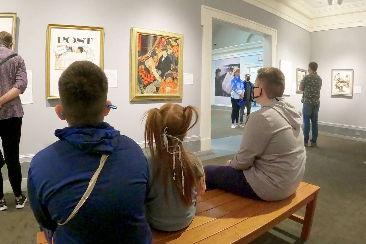 Three caucasian children wearing safety masks sit on a bench and observe paintings by Norman Rockwell indoors at the Norman Rockwell Museum.