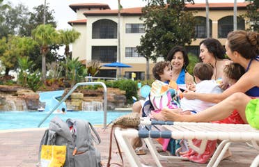 Three caucasian women wear swimwear on pool lounge chairs each holding one of three children covered in towels near a pool at Orange Lake Resort