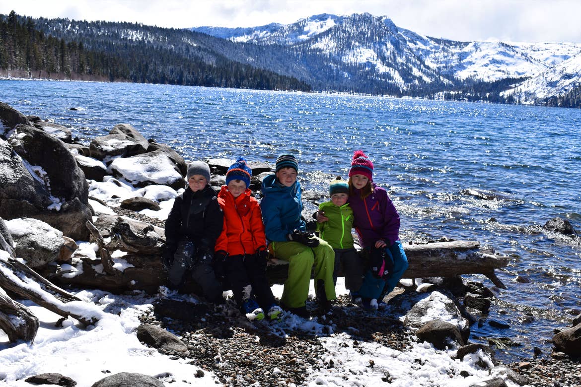 Five children sit on a snow-covered log wearing winter apparel near a lake and mountains.