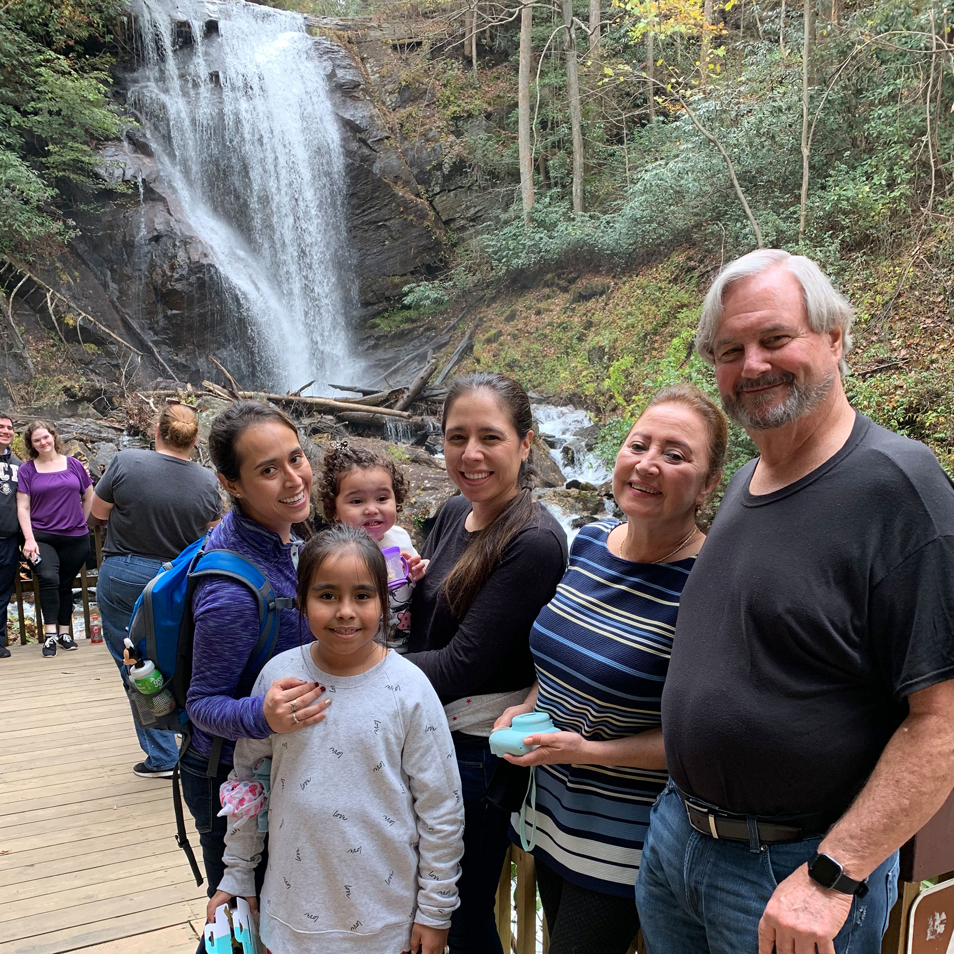 Featured author, Andrea Beltran (left), poses near a waterfall wearing a blue hiking backpack and purple pull-up sweater with her family.