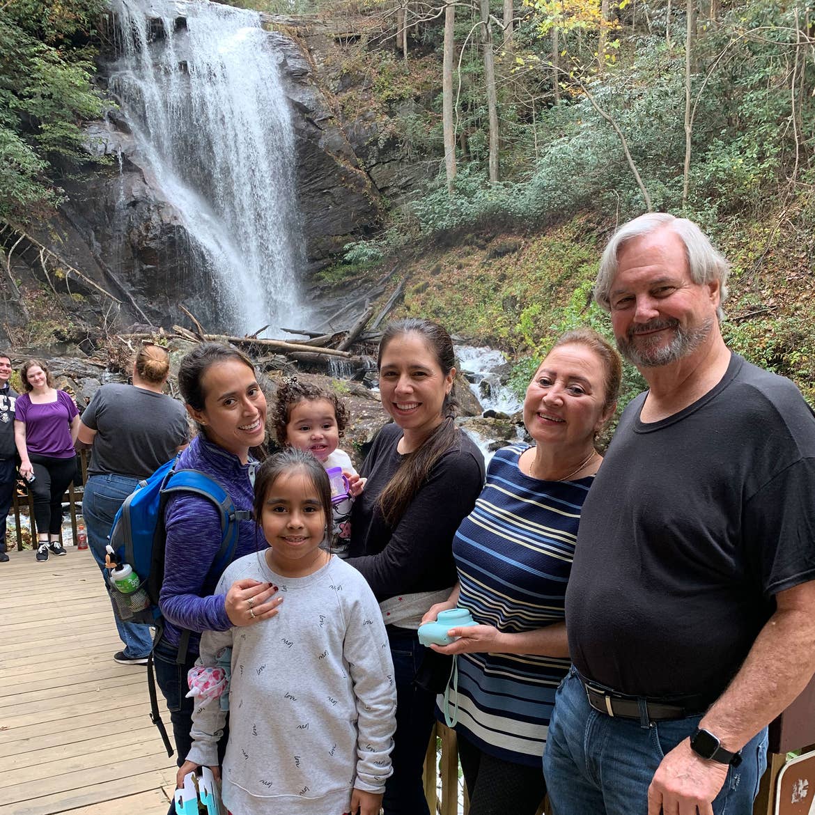 Featured author, Andrea Beltran (left), poses near a waterfall wearing a blue hiking backpack and purple pull-up sweater with her family.
