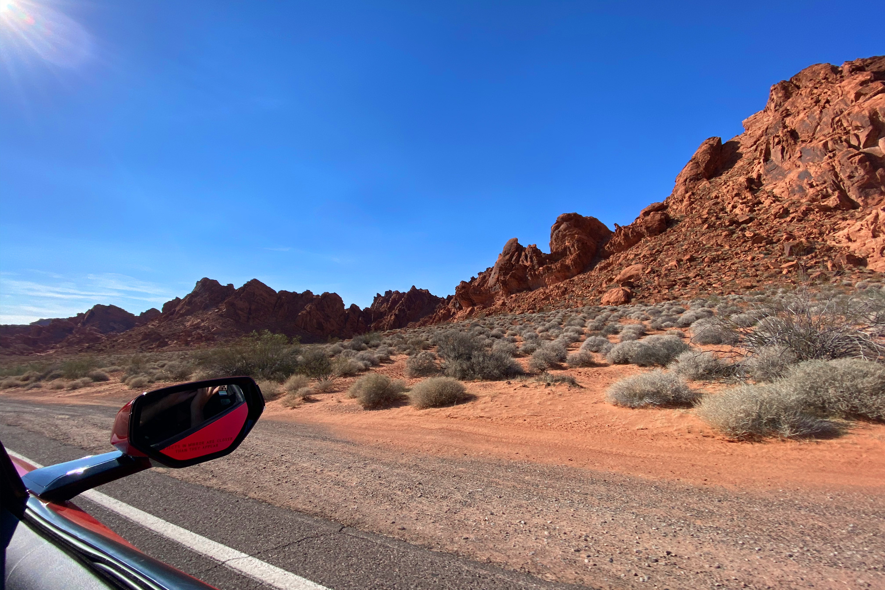 A car mirror from the passenger side encroaches the Valley of Fire rock formations while driving down a black asphalt road.