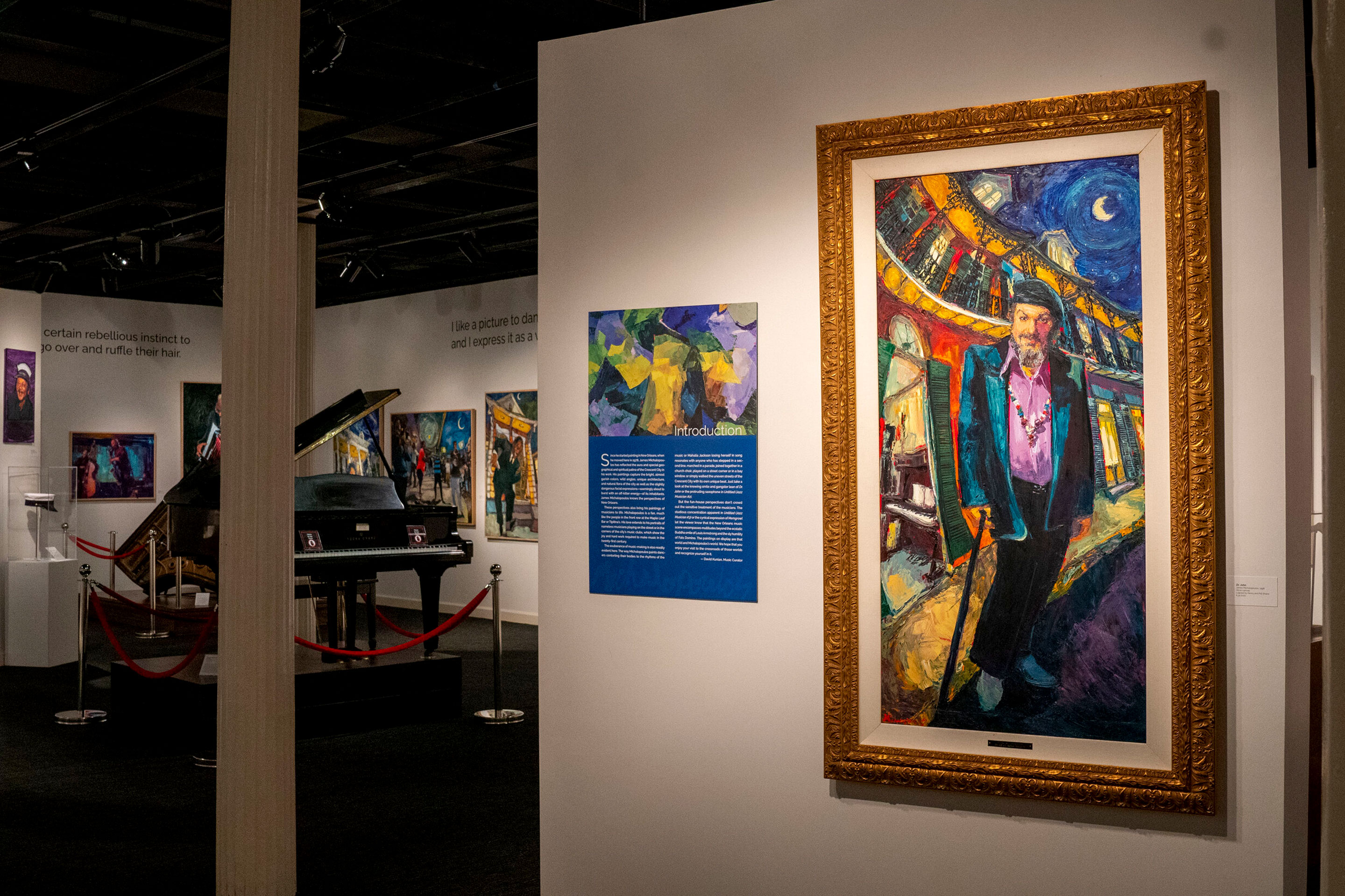 Several paintings are displayed on white walls of a museum exhibit hall with a black, baby grand piano in the center of the floor.