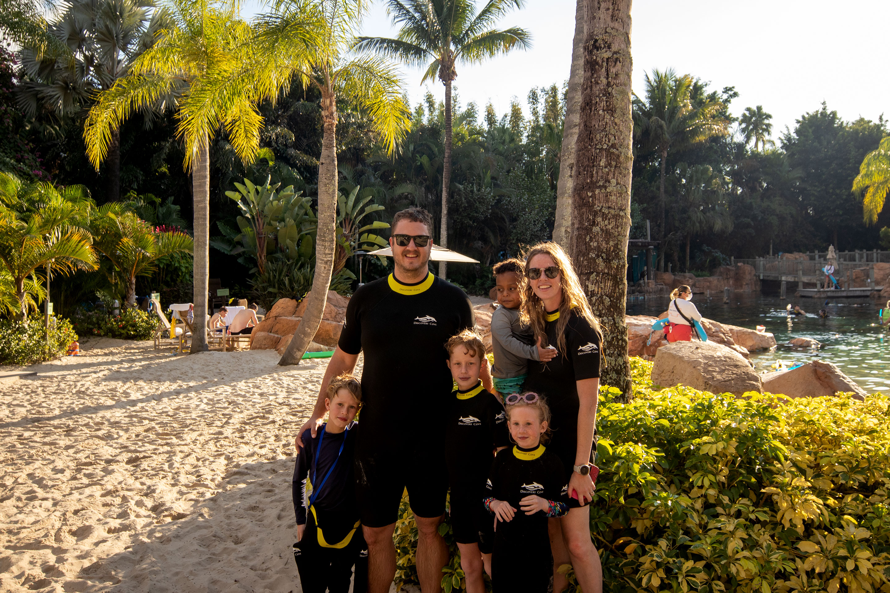 A man, woman, and four children wear black and yellow wetsuits on a sandy beach under the sun and pal trees.