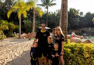 A man, woman, and four children wear black and yellow wetsuits on a sandy beach under the sun and pal trees.