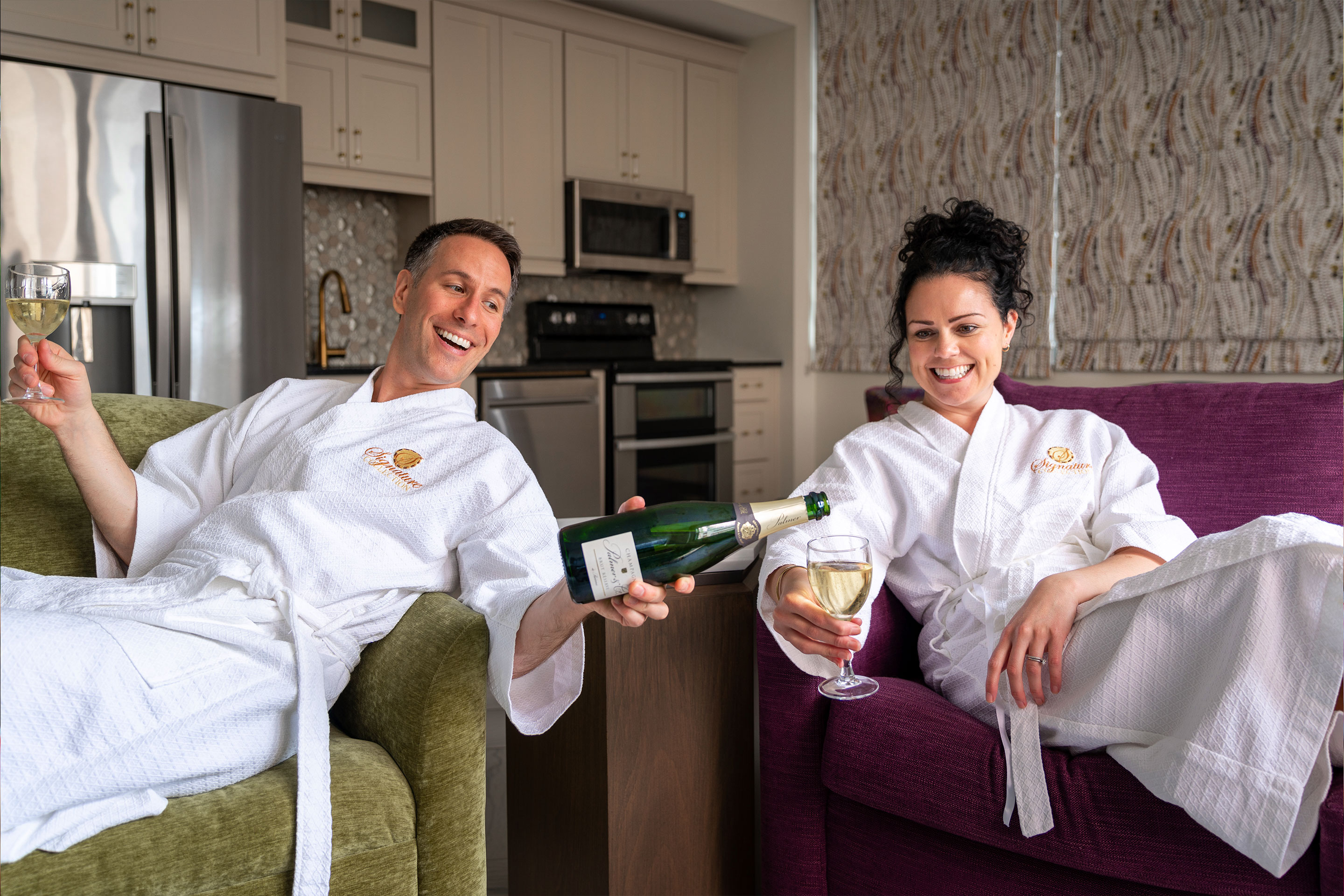 From left to right: A man and woman sitting and wearing white robes while holding a bottle and glasses of white wine.