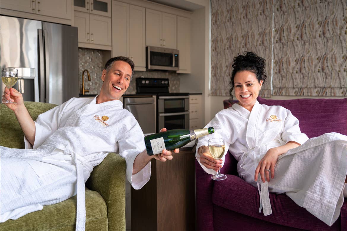From left to right: A man and woman sitting and wearing white robes while holding a bottle and glasses of white wine.