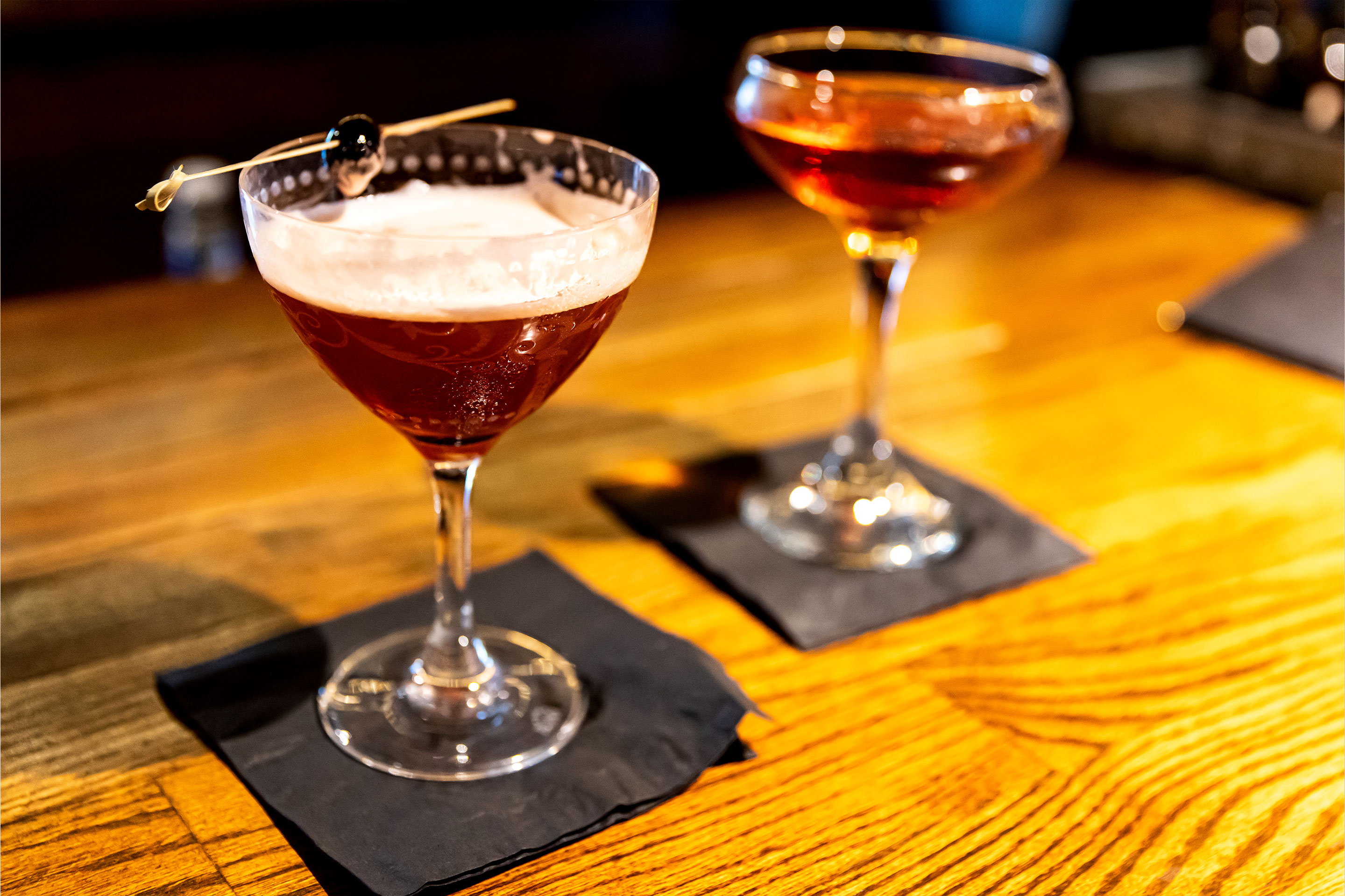 Two cocktail glasses are placed on black napkins on a wooden bar top.