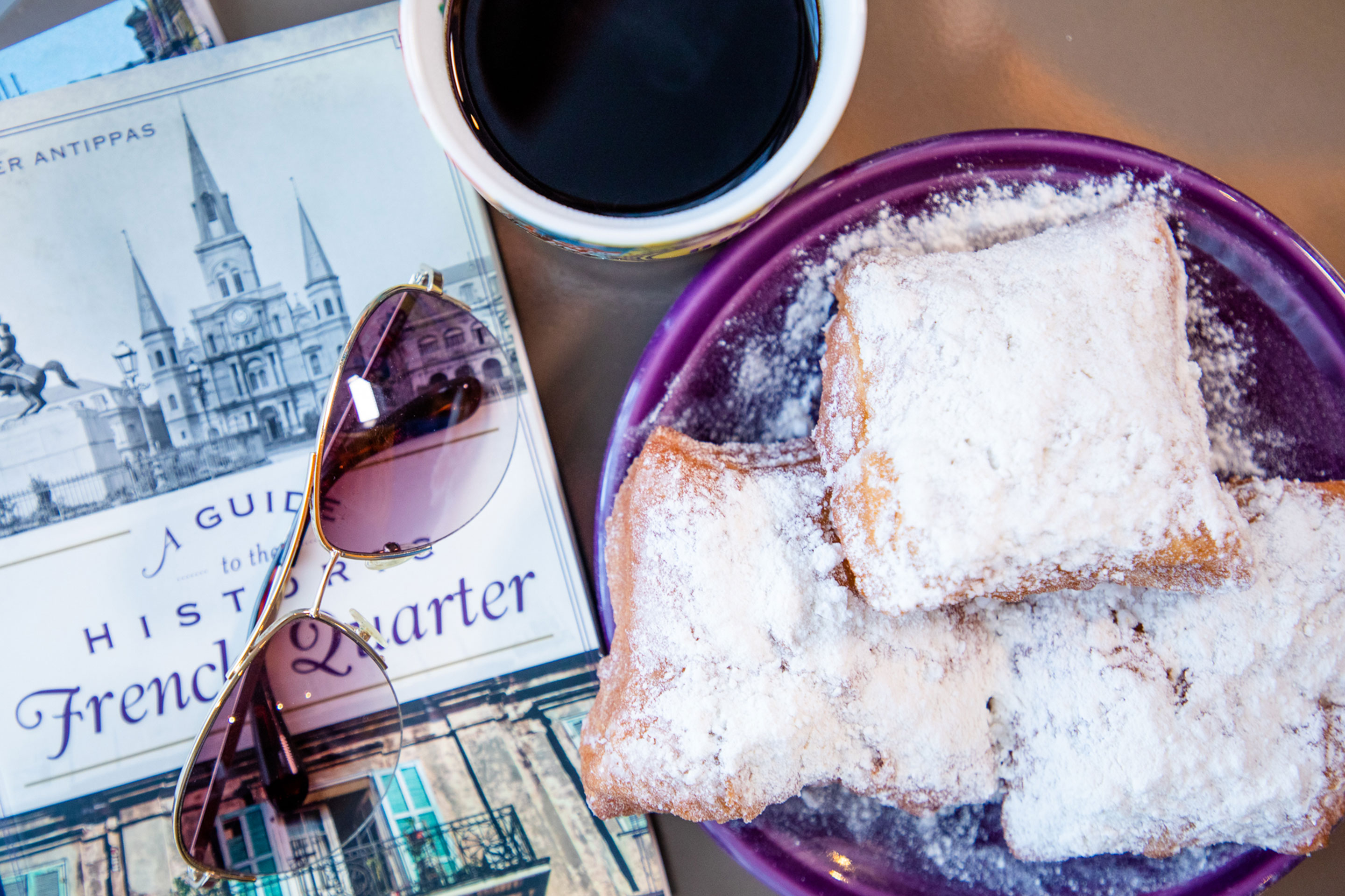 A tabletop with three beignets on a purple plate surrounded by a pair of sunglasses, a cup of chicory coffee in a white mug, and a book with the title, 'A Guide to the Historic French Quarter.'
