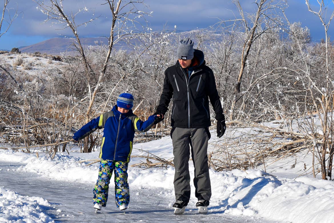 Featured Contributor, Jessica Averett's husband (right) wears a black winter coat, snow pants, and white ice skates as he helps his son (right) wearing blue and lime winter jacket, snow pants, and white ice skates outside.