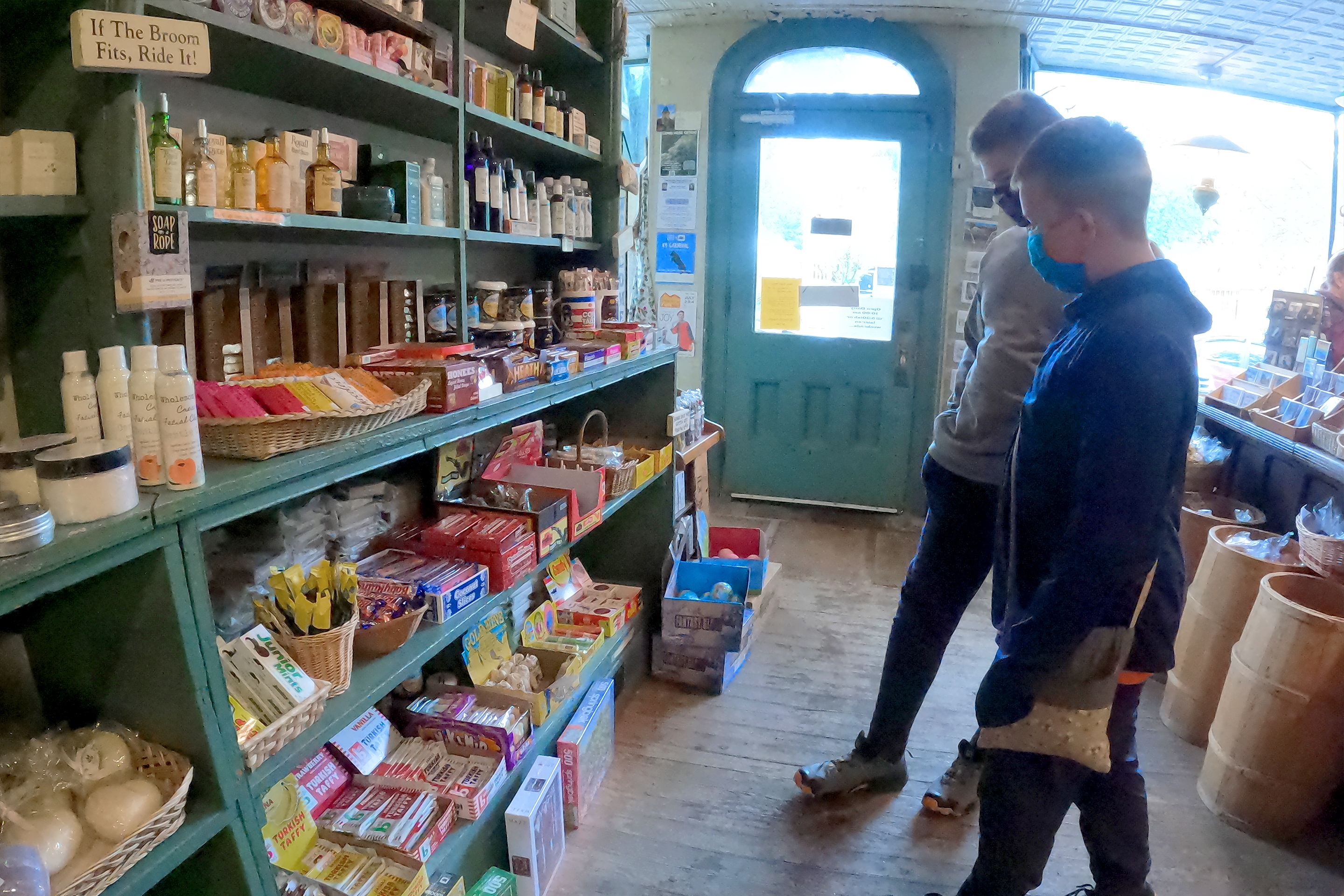 Two caucasian tween boys wearing long pants, hoodies and safety masks stand inside a candy store looking at various treats on the wall.