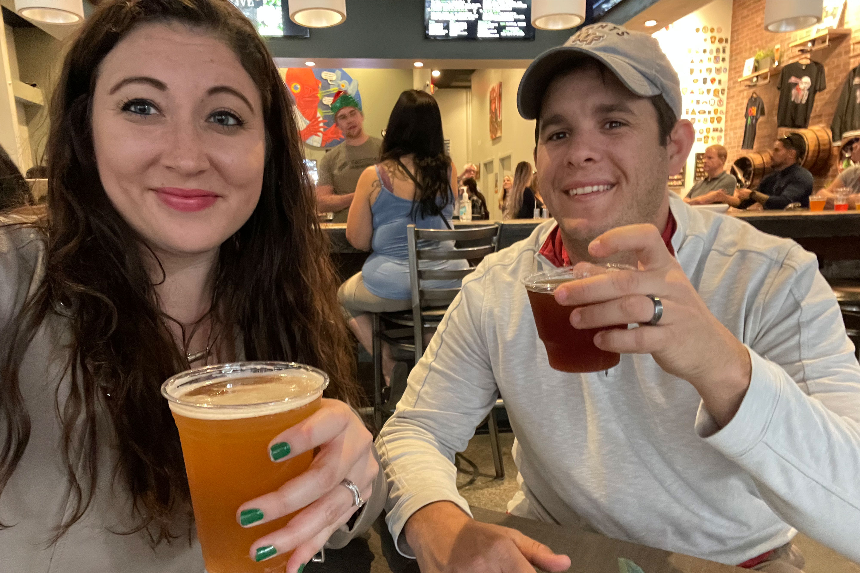 A woman in a tan top and a man in a white pullover and backward cap hold up their glasses of beer for a toast.