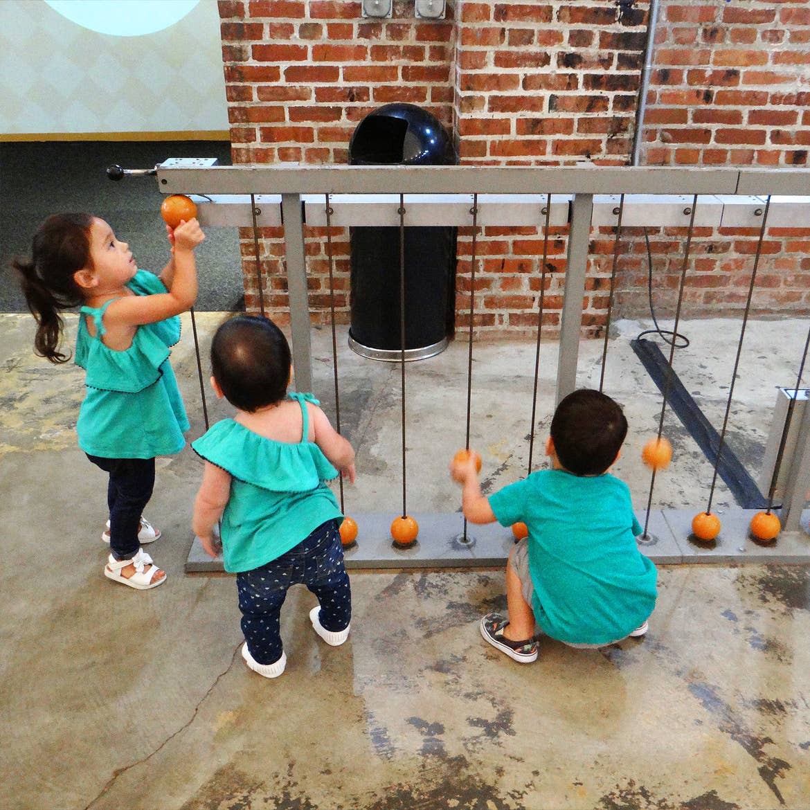 Three toddlers in matching denim and teal shirts interact with a display indoors.
