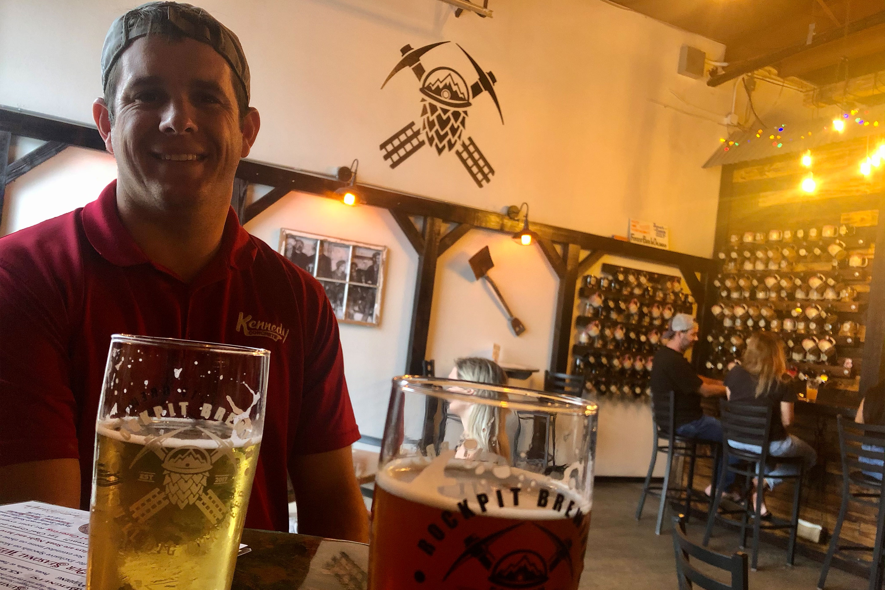 A man in a red polo and backwards cap sits at a wooden bar top with two glasses of beer.