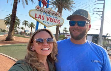 A caucasian woman wearing sunglasses (left) poses near a caucasian man wearing sunglasses and a hat (right) in front of the 'Welcome to Las Vegas Nevada' marquee sign.