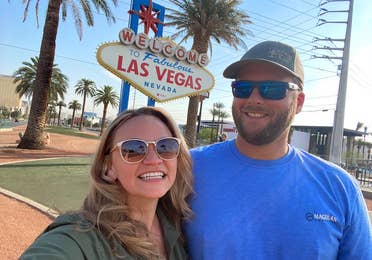 A caucasian woman wearing sunglasses (left) poses near a caucasian man wearing sunglasses and a hat (right) in front of the 'Welcome to Las Vegas Nevada' marquee sign.