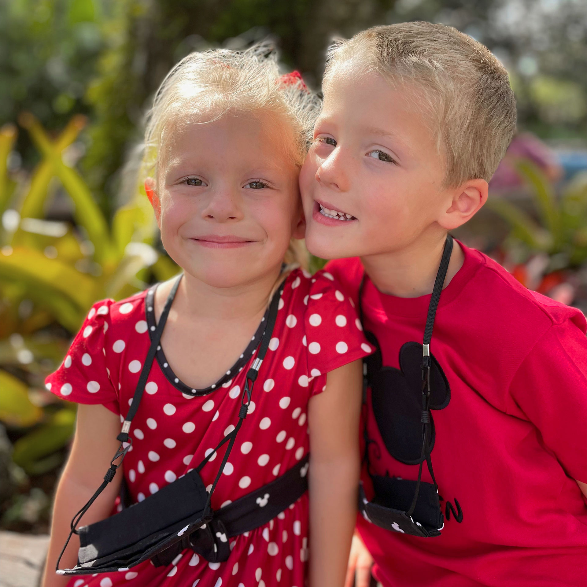 A girl wearing a red and white polka-dot dress and a boy wearing a red t-shirt wear black masks hanging from lanyards outdoors.