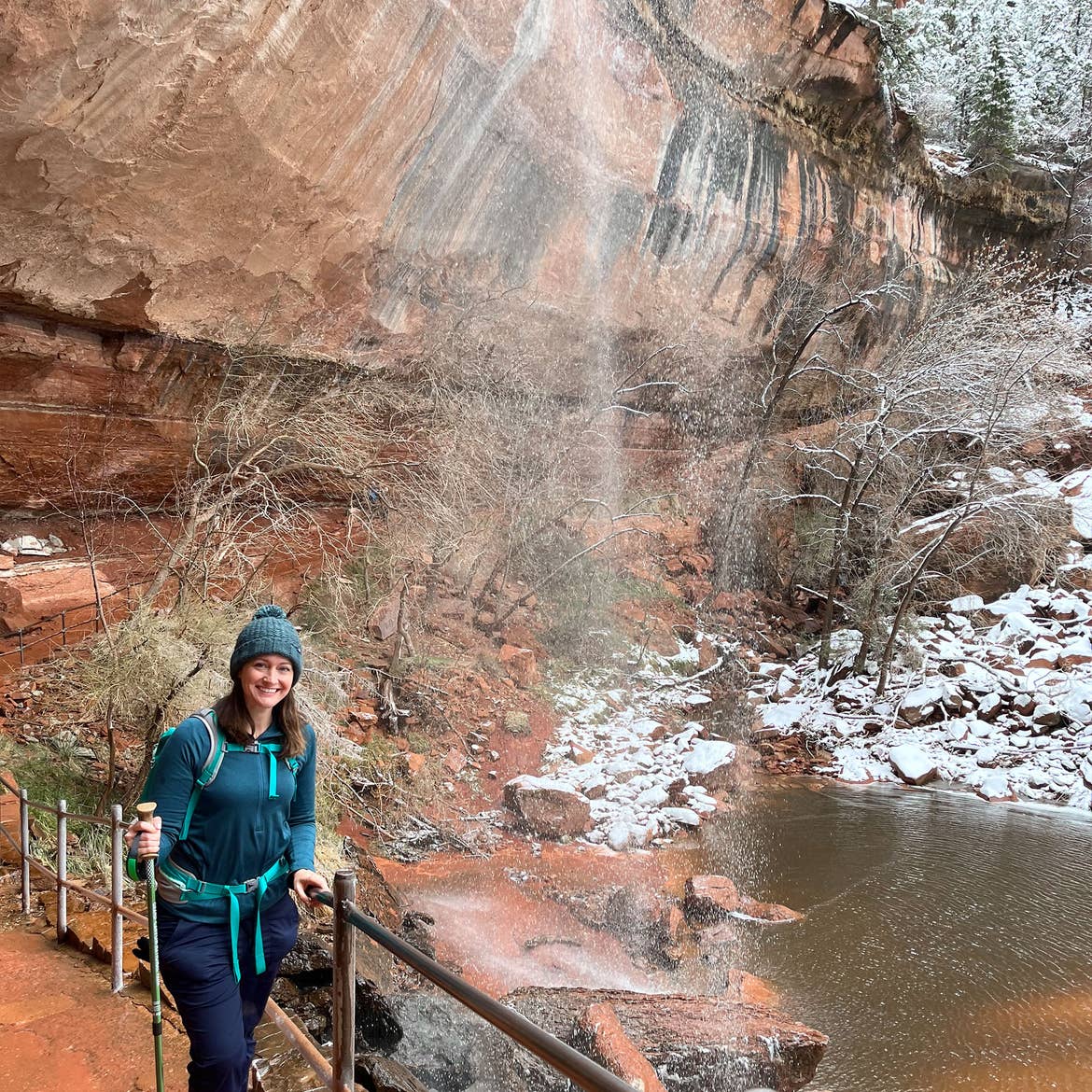 A woman in an aqua pullover, knitted cap and holding walking sticks stands near a waterfall.