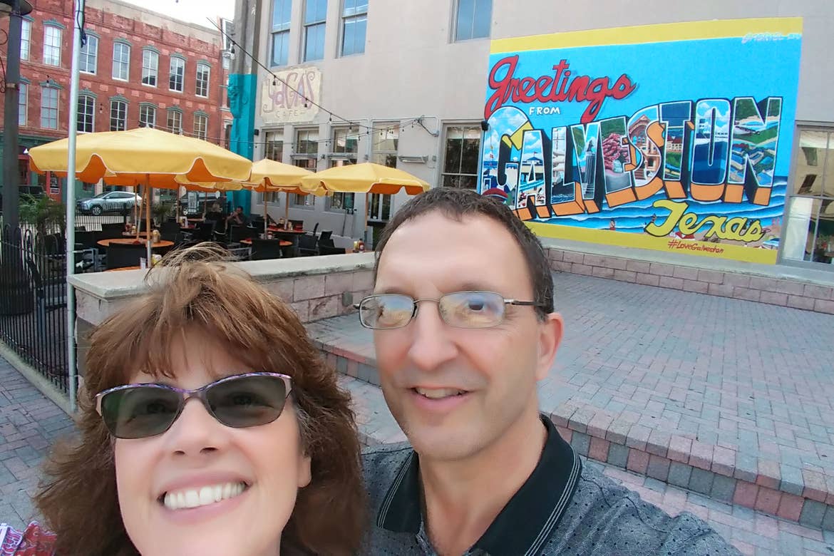 A woman in sunglasses and a man in reading glasses stand near a mural that reads, 'Greetings from Galveston, Texas' near a brick-paved walkway.