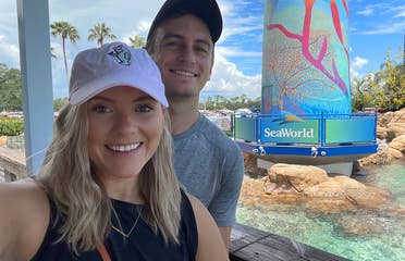 A Caucasian female wearing a pink baseball cap and black tank top stands near the lighthouse in SeaWorld Orlando.