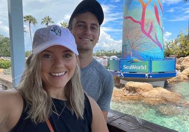A Caucasian female wearing a pink baseball cap and black tank top stands near the lighthouse in SeaWorld Orlando.