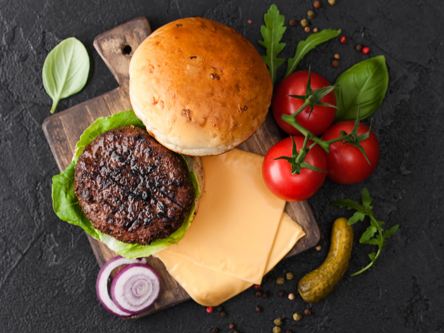 Burger preparations with bun, patty, cheese, tomatoes, pickle and onion on cutting board.