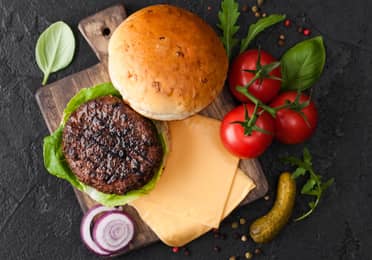 Burger preparations with bun, patty, cheese, tomatoes, pickle and onion on cutting board.