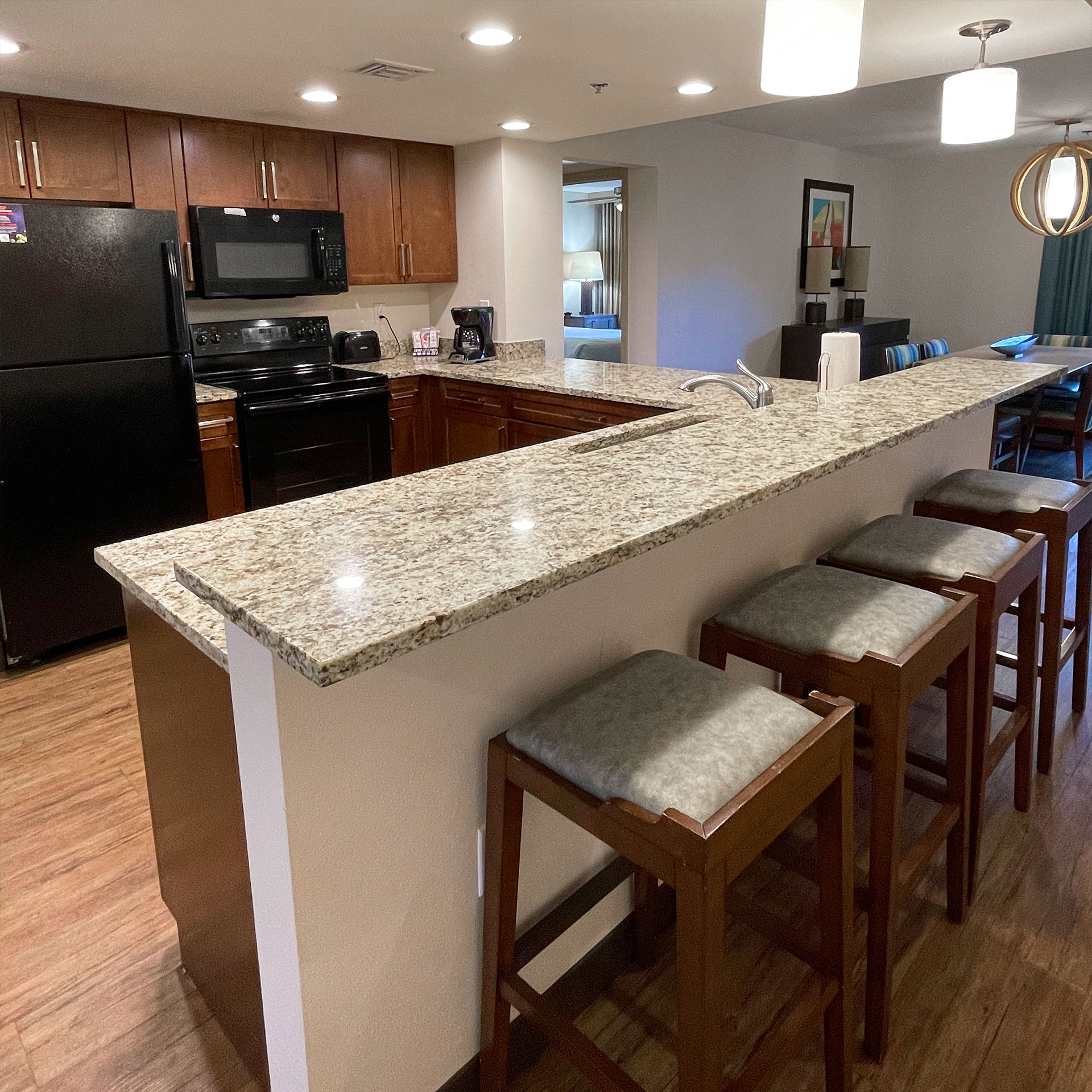 A kitchen interior space with black appliances and light, granite countertops overlooking into the living room area.