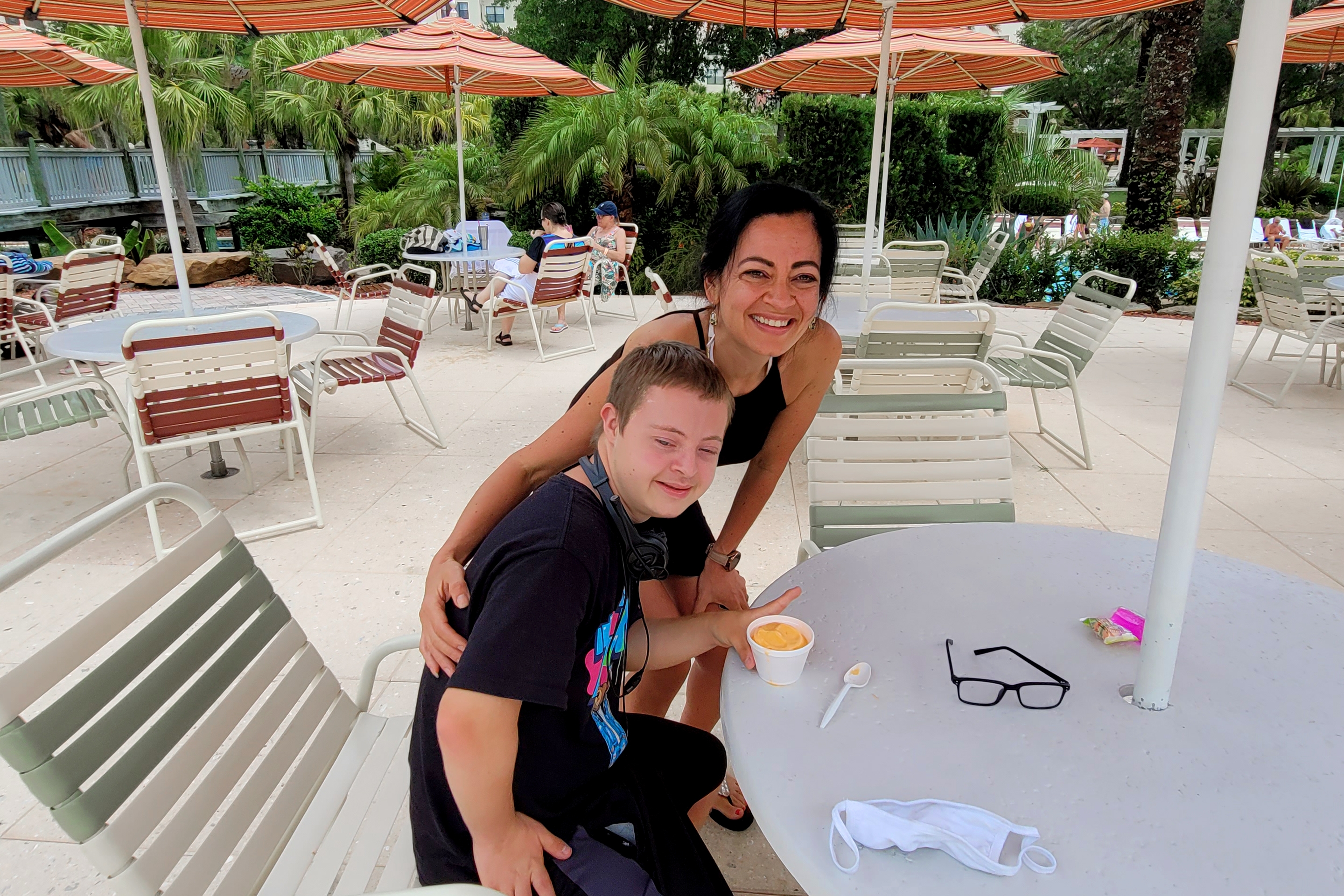 A woman (back) stands behind a young boy (front) eating a cup of ice cream.