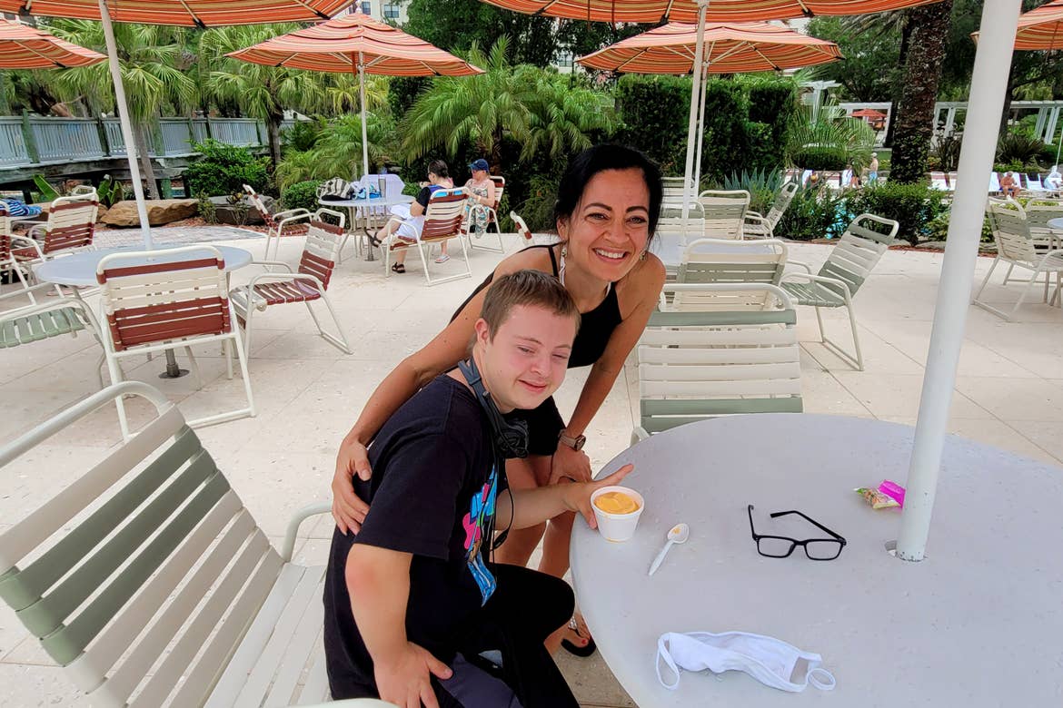 A woman (back) stands behind a young boy (front) eating a cup of ice cream.