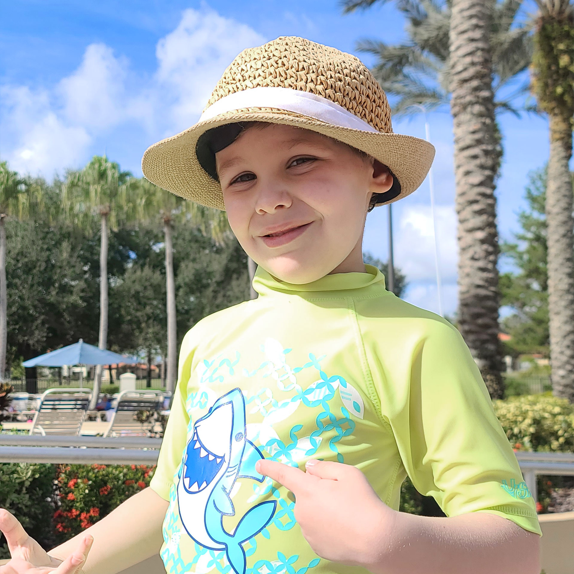 A boy in a straw hat and yellow rash guard stands under palm trees on a pool deck.