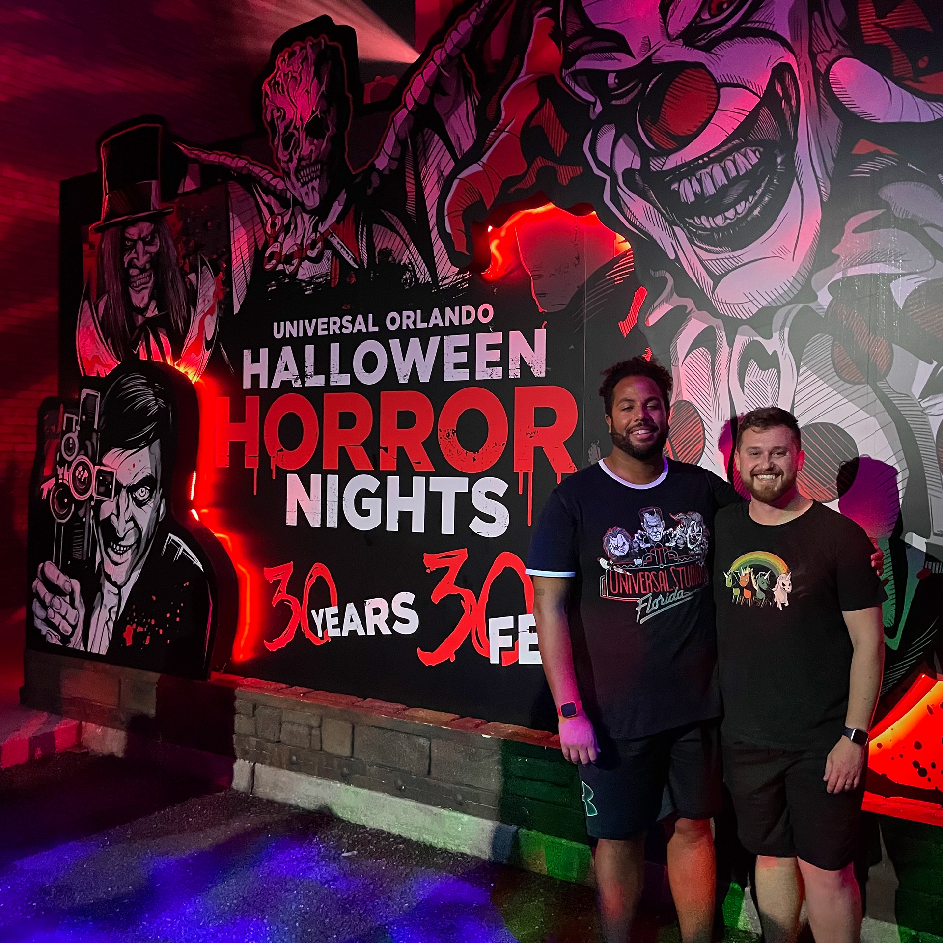 Two men wearing black t-shirts and shorts stand in front of a mural at night.