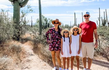 A woman in a floral dress, two young girls wearing white dresses, and a man in a red shirt and khaki shorts wear hats and sunglasses in a desert surrounded by cacti under a blue, cloudy sky.