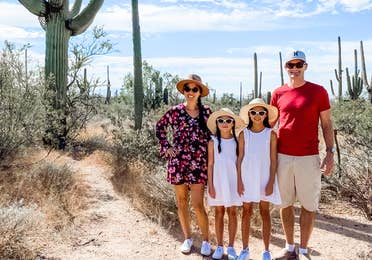 A woman in a floral dress, two young girls wearing white dresses, and a man in a red shirt and khaki shorts wear hats and sunglasses in a desert surrounded by cacti under a blue, cloudy sky.