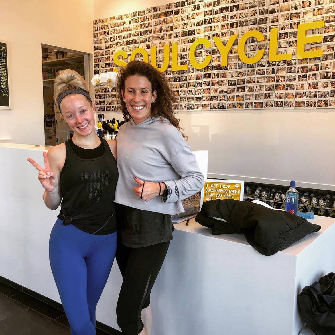 Co-author, Molly (left), wears a black tank and blue leggings while posing with a friend near a SoulCycle entrance.
