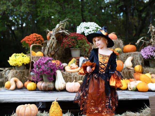 Child dressed in a witch Halloween costume standing outside by hay bales and pumpkins.