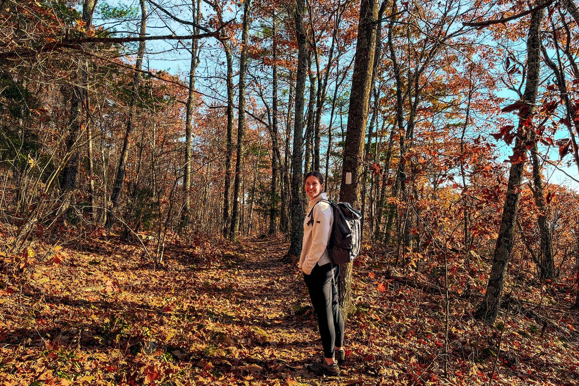 A woman wearing a white pullover with black pants and a hiking backpack stands on a trail path surrounded by trees with fall foliage.