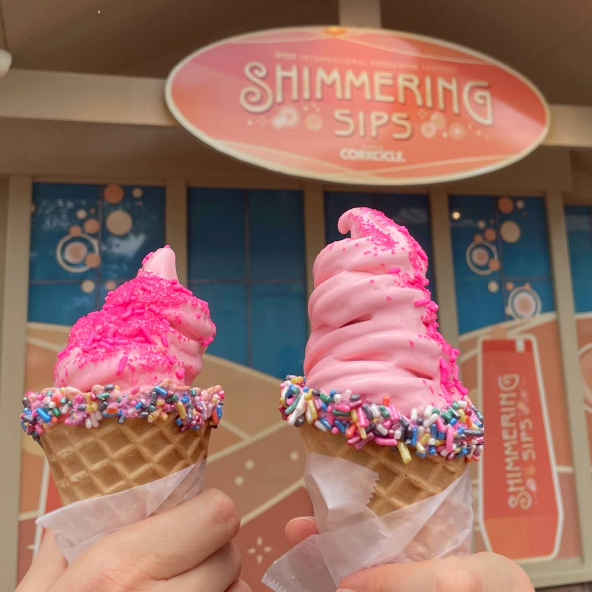 Two hands hold two pink soft-serves in sprinkled waffle cones topped with pink sprinkles near the Corkcicle Shimmering Sips kiosk at Epcot.