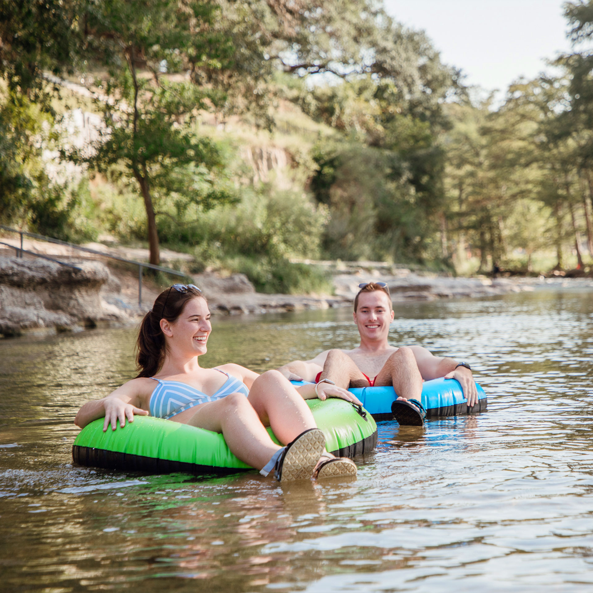 A woman in a blue and white bikini floats in a green inner tube next to a man in red swim trunks floating in a blue inner tube down a stream.