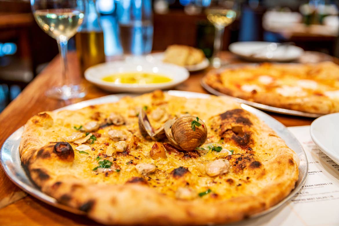 A clam Pizza placed on a dining table surrounded by glasses of white wine.