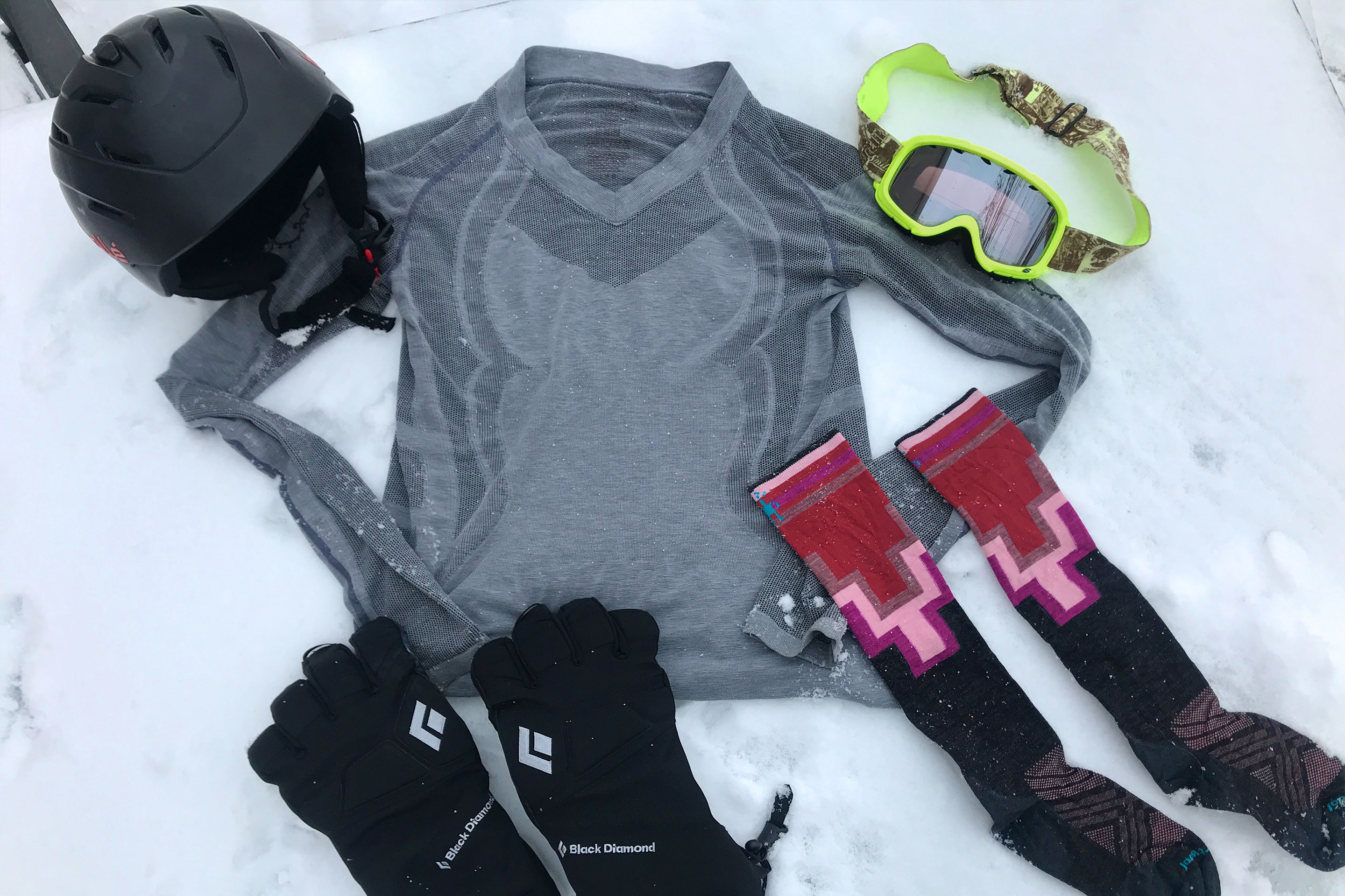 (From top-left to bottom-right) A black ski helmet, grey, long-sleeved shirt, neon yellow snow goggles, black gloves, and multi-colored high socks are place on snow.