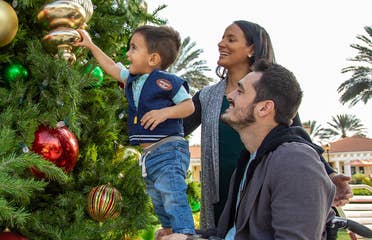 Author, Danny Pitaluga (right) sits in his black wheelchair wearing a charcoal hoodie as his wife, Val (middle), and his son, Joey (left), stands on Danny's lap to decorate a Christmas tree outdoors.