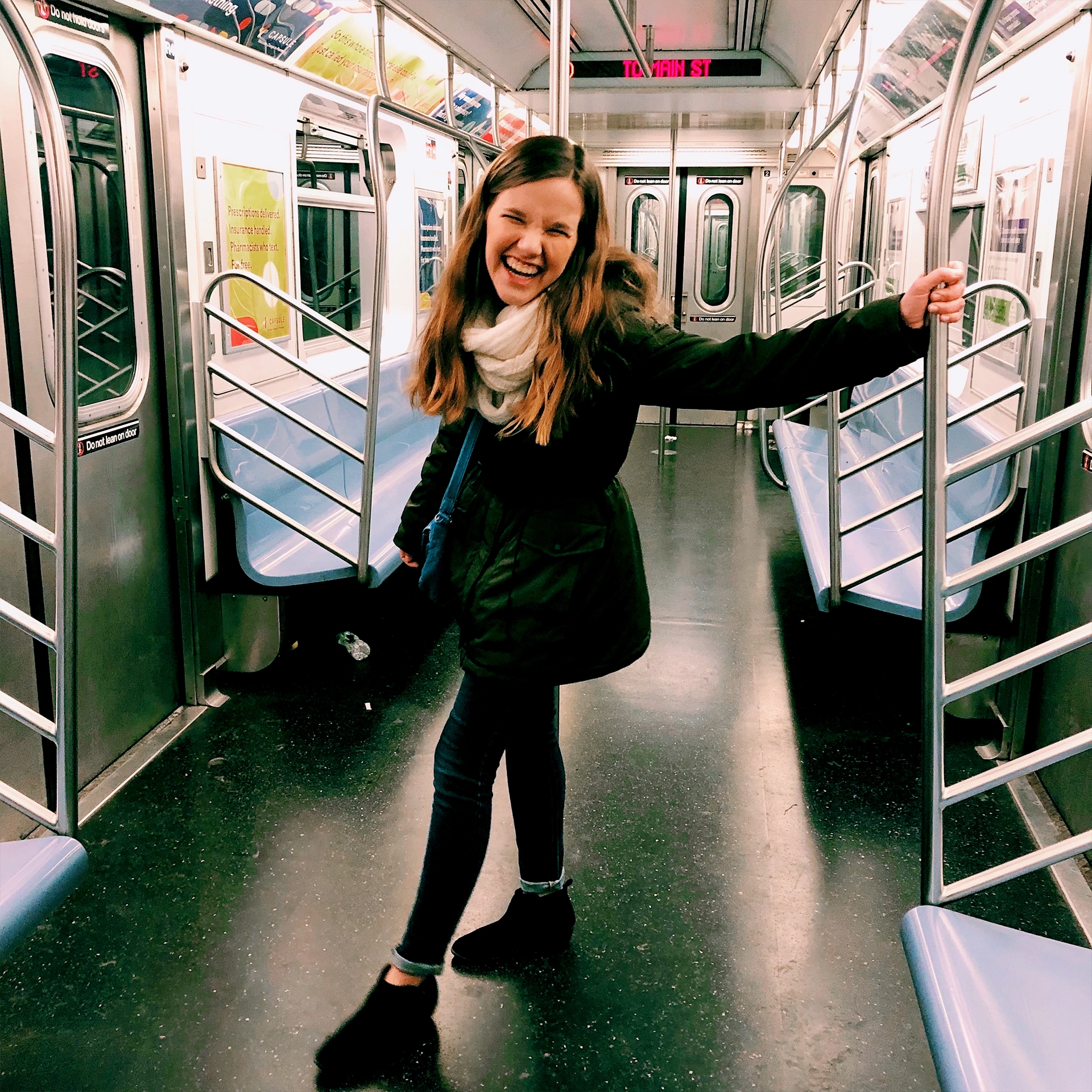 A caucasian woman with long ash-blonde hair wears a white scarf and green winter jacket while standing in a subway in New York City.